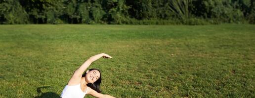 Young woman does yoga on lawn in park, stretching on fitness mat, wellbeing concept photo