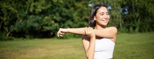 retrato de joven aptitud mujer extensión su brazos, calentar antes de formación sesión, deporte evento en parque, trotar y ejercitándose foto