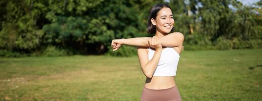 Asian woman stretching her arms, doing fitness workout in park, smiling pleased, warming up before jogging on fresh air in daytime photo