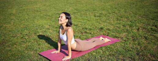 Smiling young sportswoman stretches on rubber mat in park, does yoga asana exercises, workout on fresh air in fitness clothing photo