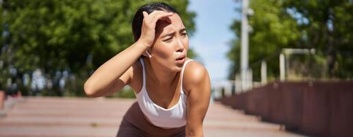 Tired young female runner, asian girl taking break during workout, stop jogging, panting while breathing, running in park photo