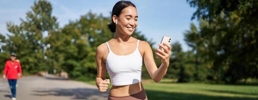 Fitness woman with water bottle and smartphone, jogging in park and smiling, looking at her mobile phone app, checking sport application photo