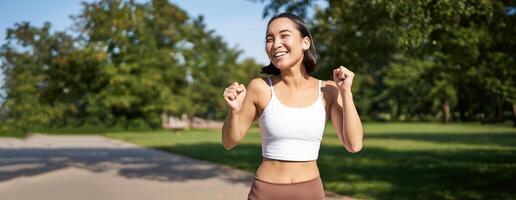 Happy fitness girl achieve goal, finish marathon, running with hands up, celebrating victory while jogging, triumphing in park photo