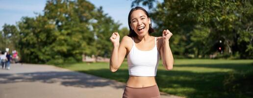 Happy fitness girl achieve goal, finish marathon, running with hands up, celebrating victory while jogging, triumphing in park photo