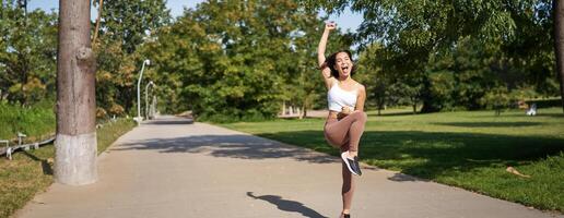 Happy asian girl celebrating victory, saying yes, finish running, workout in park with excitement and joy photo