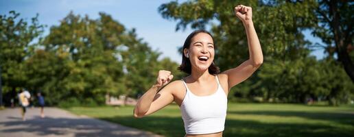 Happy fitness girl achieve goal, finish marathon, running with hands up, celebrating victory while jogging, triumphing in park photo