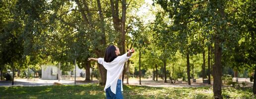 despreocupado asiático niña baile, sensación felicidad y alegría, disfrutando el Dom en verano día, caminando en parque con verde arboles foto