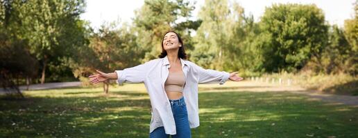 Carefree asian girl laughing and dancing in park, enjoying summer sunny day, raising hands up and breathing fresh air photo