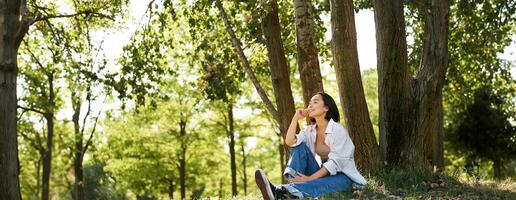 Relaxed young woman, resting near tree, sitting in park on lawn under shade, smiling and looking happy, walking outdoors on fresh air photo