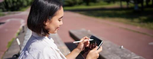 Portrait of young asian woman drawing on fresh air in park, sitting with graphic tablet and digital pen, smiling happy photo