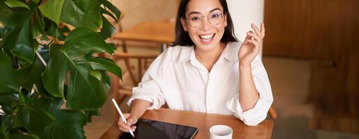 Young asian creative girl, sitting with digital tablet, drawing with graphic pen and smiling, doodling, drinking coffee in cafe photo