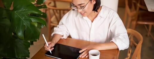 Young asian creative girl, sitting with digital tablet, drawing with graphic pen and smiling, doodling, drinking coffee in cafe photo