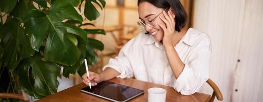 Young asian creative girl, sitting with digital tablet, drawing with graphic pen and smiling, doodling, drinking coffee in cafe photo