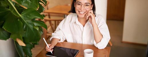 Young asian creative girl, sitting with digital tablet, drawing with graphic pen and smiling, doodling, drinking coffee in cafe photo