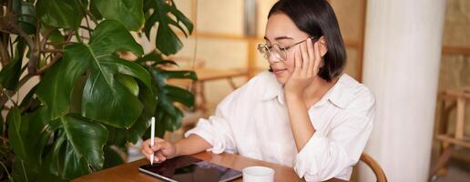 Young artist drawing on digital tablet, using graphic pen, drinking coffee in cafe and scatching smth photo