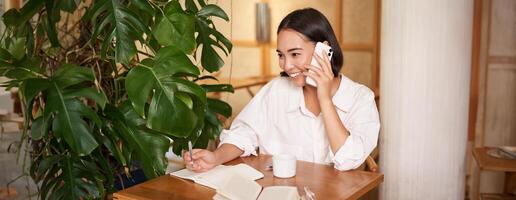 Working woman answer phone call in cafe, writing down, making notes while having conversation on telephone photo