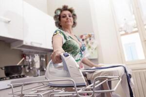 beautiful mature woman with curlers ironing clothes at home photo