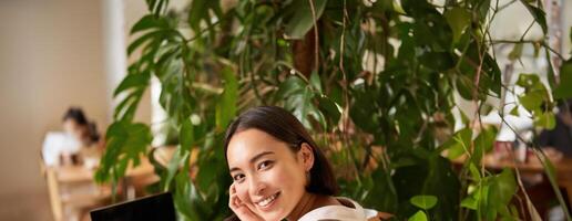 Trendy young woman sitting in cafe and smiling at camera, drinking coffee and using laptop, working remotely, studying or browsing internet photo