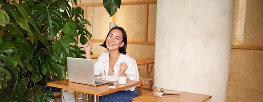 contento joven asiático mujer victorioso en computadora portátil, recibir bueno noticias, lograr objetivo a trabajar, triunfando y sonriente complacido, sentado en café foto