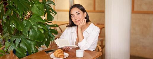 Dreamy young smiling asian woman reading book, sitting in cafe, eating croissant and drinking coffee in cozy interior photo