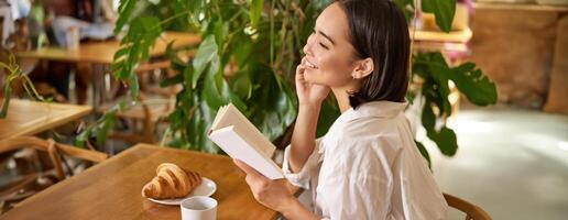 hermosa joven asiático mujer con un libro en manos, sentado en cafetería, Bebiendo café y comiendo cuerno, sonriente, mirando misterioso foto