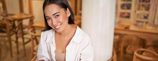 Vertical shot of young smiling woman with book, reading in cafe, enjoying morning breakfast coffee and croissant photo