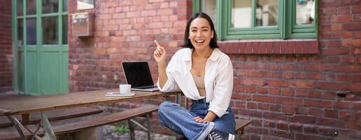 Enthusiastic asian woman, pointing at upper right corner, showing advertisement, news on banner, looking impressed and intrigued by smth photo