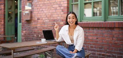stylish modern asian girl with laptop, sitting in cafe, looking amazed and pointing at upper right corner banner, showing info advertisement photo