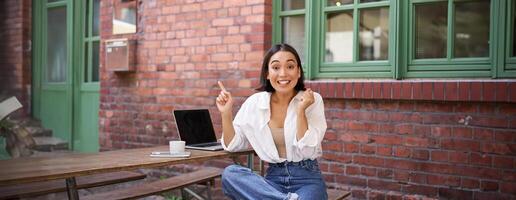 Stylish urban girl, asian woman with laptop sits in cafe, points at upper right corner banner, shows copy space with surprised face expression photo