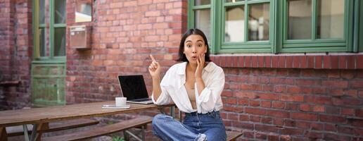 Stylish urban girl, asian woman with laptop sits in cafe, points at upper right corner banner, shows copy space with surprised face expression photo