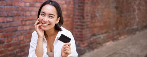 Beautiful young asian woman with credit card, sitting near laptop and smiling, paying bills, shops online, order smth on computer photo