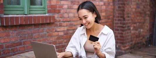 Smiling asian woman sitting with laptop, paying by credit card for online shopping, sending her bank account details, sitting in cafe photo