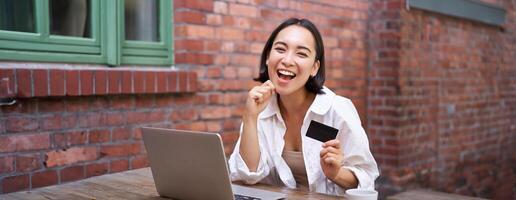 contento joven asiático mujer sentado cerca computadora portátil, participación crédito tarjeta, pago facturas, compras en línea sin contacto, sonriente a cámara foto