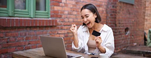 Happy young asian woman sitting near laptop, holding credit card, paying bills, shopping online contactless, smiling at camera photo