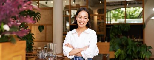 Young asian woman, cafe owner or manager, standing confident with arms crossed on hands, smiling and looking at camera photo