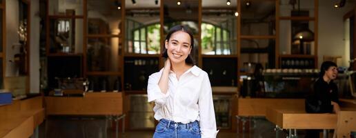 Portrait of cute and shy young asian woman, standing in front of cafe entrance, touching her neck and smiling, introducing her business photo