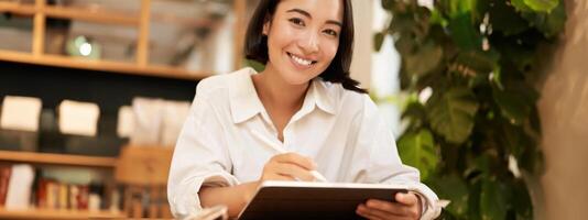 Portrait of asian girl drawing on digital tablet, scatching and doodling with graphic pen, sitting in cafe and smiling photo