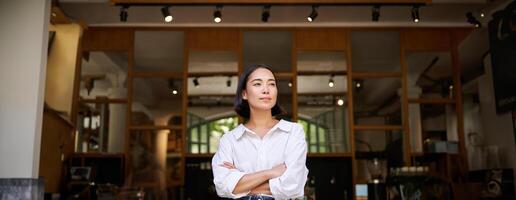 Portrait of confident young asian woman, business owner, cross arms on chest, looking pleased, standing in front of cafe photo