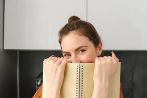 Portrait of young modern woman, 25 years old, standing in the kitchen with notebook, reading notes, student revising for exam photo