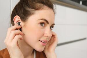 Portrait of young smiling woman listens to music in her black wireless earphones, using headphones to enjoy sound quality of song, sits on kitchen floor photo