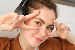 Close up portrait of woman in headphones, shows peace, v-signs near eyes, smiles and looks happy, sits on floor at home photo