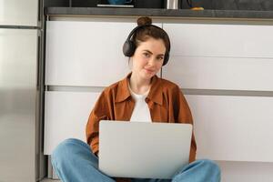 Young woman, student doing homework on laptop, sitting on floor, listening to music in headphones and working from home photo