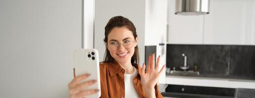 Portrait of stylish beautiful woman at home, video chats on smartphone, connects to online meeting from her living room, lifestyle social media blogger records a video on mobile phone photo