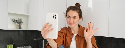 Portrait of young cheerful woman laughing and smiling during phone call, video chats with friend, sits on kitchen counter and talks to someone using smartphone app photo