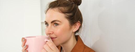 acogedor momentos contento joven mujer se sienta en cocina con taza de recién elaborada café, sonriente y mirando aparte. niña bebidas té a hogar foto