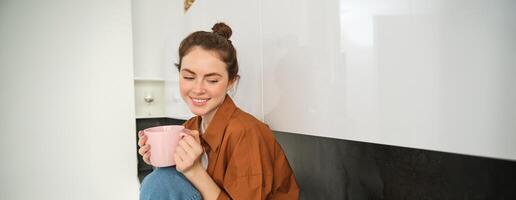 retrato de joven mujer con taza de café, se sienta en cocina y bebidas aromático bebida a hogar, sostiene té jarra foto