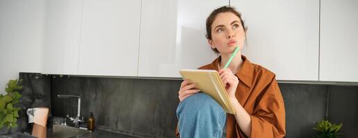 Image of young creative woman, artist drawing sketches in her notebook, sitting on kitchen counter at home with thoughtful face photo