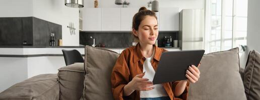 Portrait of female model reading on digital tablet e-book, watching tv series on her gadget application, sitting in living room at home photo