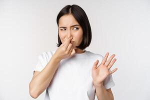 Asian girl looks disgusted, rejecting product with bad smell, shut nose from aversion and cringe, standing against white background photo