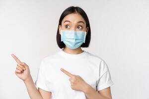 Young korean woman in medical face mask pointing fingers left and looking at logo, showing advertisement or banner, standing over white background photo